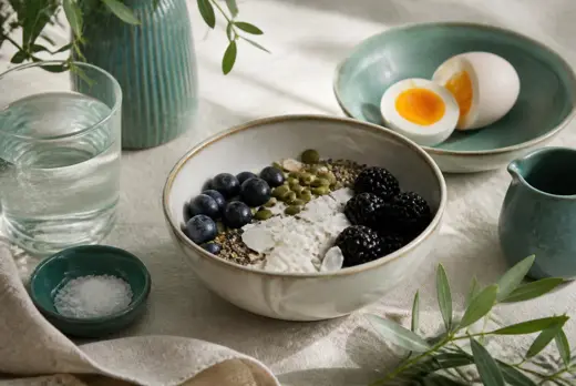 A calm breakfast of eggs, berries, seeds and water on cream linen in soft daylight